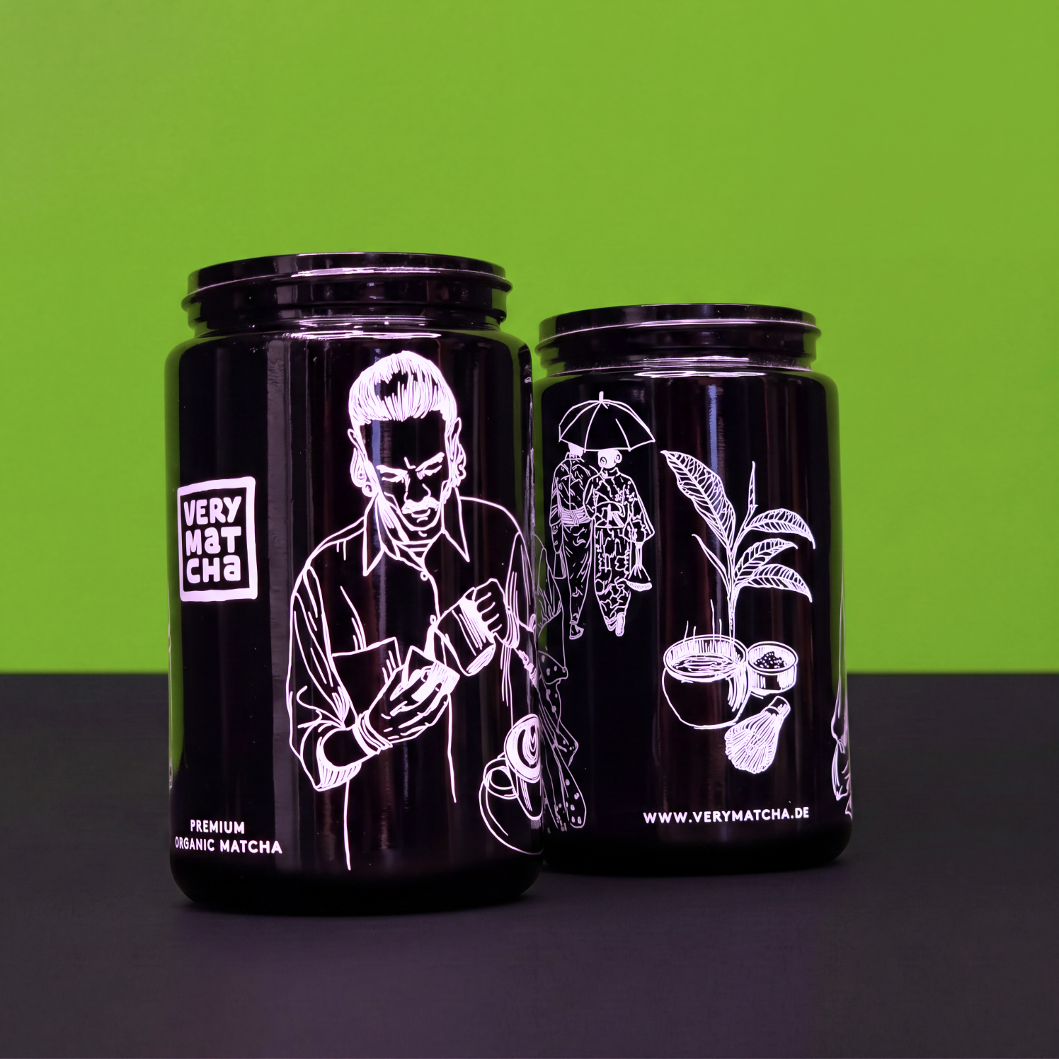 Zwei schwarze Matcha-Gläser mit weißem Siebdruck vor grünem Hintergrund